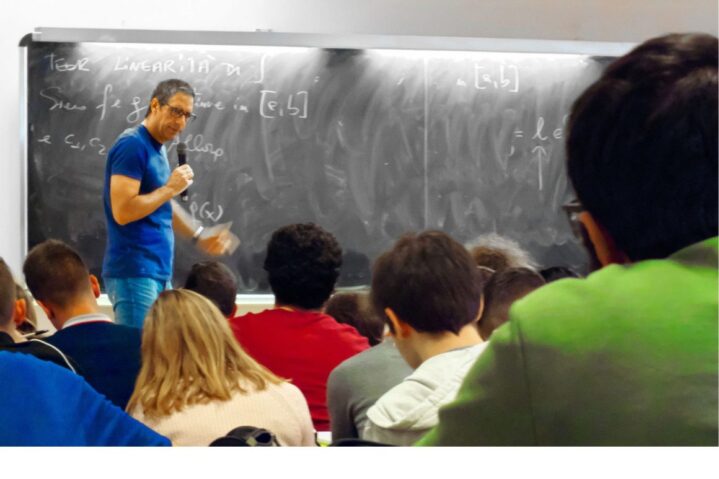 An image of a classroom full of students where a lecture is taking place. A professor holding a microphone and standing in front of the class next to a black board.