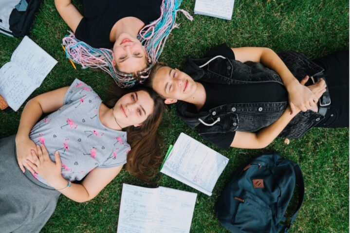 Three students are lying on the grass in a circle with their heads close together, smiling and relaxed. Notebooks and a backpack are scattered nearby.