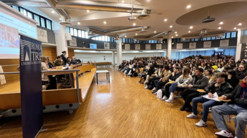 A large modern lecture hall filled with students seated, facing a panel of speakers at the front during an event or class.
