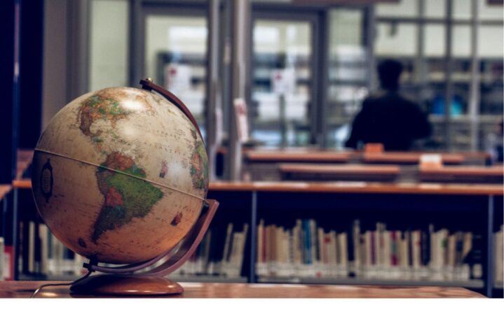 A globe is placed on a library table, with books on shelves and a person in the background, slightly out of focus - quiet study or research environment.