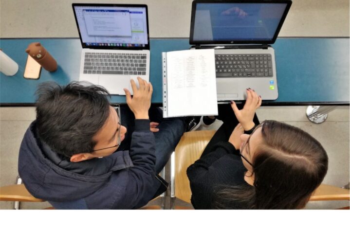 Two students are sitting side by side at a desk, studying together and working on laptops with a notebook open between them.