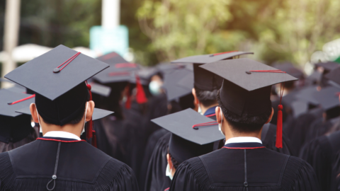 Group of students wearing black graduation gowns and caps with red tassels, standing together outdoors, facing away from the camera, seen from behind. A Picture symbolizing a graduation ceremony.
